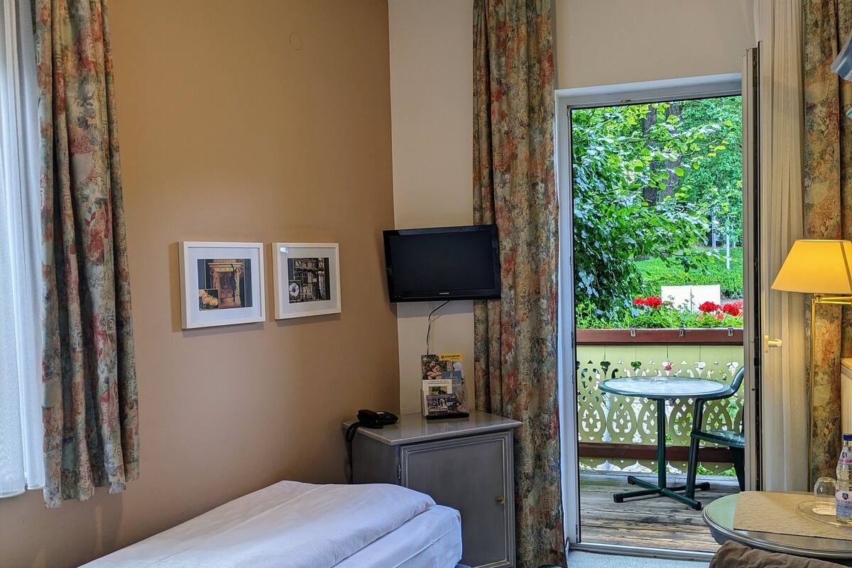 A hotel room with a single bed, a desk with a computer, a TV, and a balcony with a table and chair.