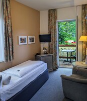 A hotel room with a single bed, a desk with a computer, a TV, and a balcony with a table and chair.