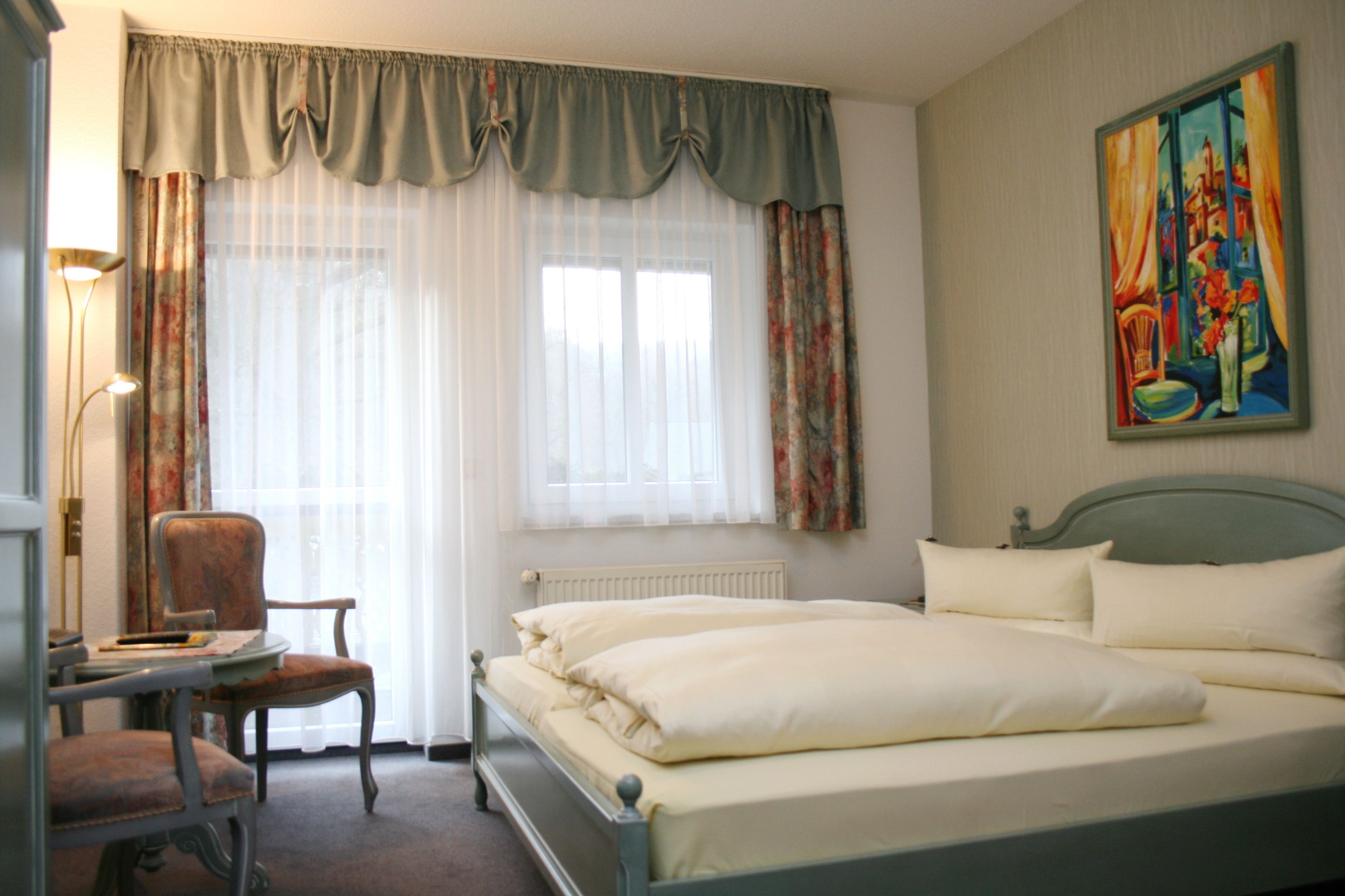 A hotel room with a bed, a chair, a desk, a radiator, and a painting on the wall.
