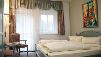 A hotel room with a bed, a chair, a desk, a radiator, and a painting on the wall.