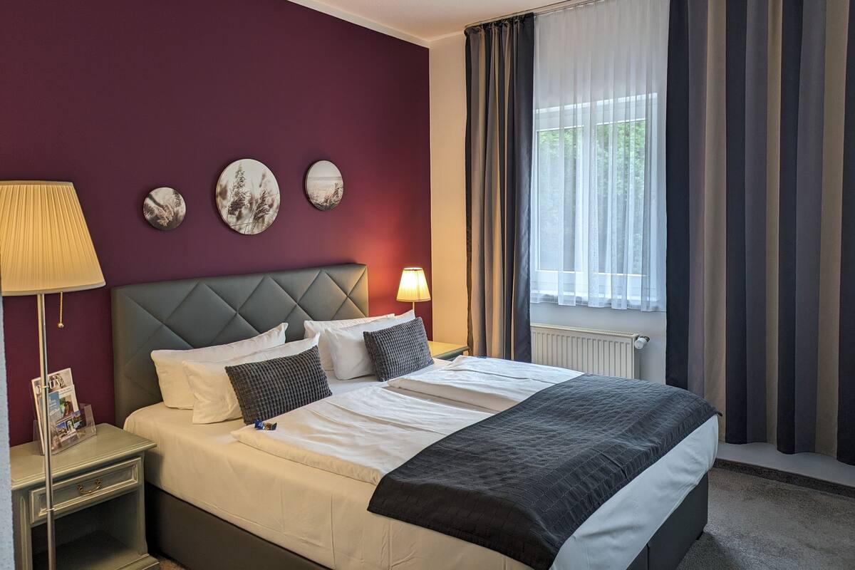 A hotel room with a large bed, two bedside tables, a nightstand, a window with curtains, and wall art.