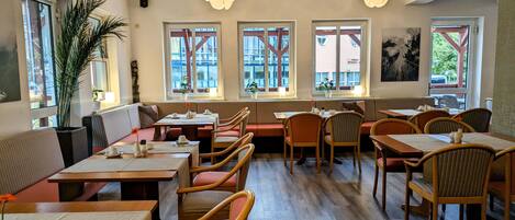 A restaurant with wooden tables and chairs, large windows with views of buildings, and hanging branches above the tables.