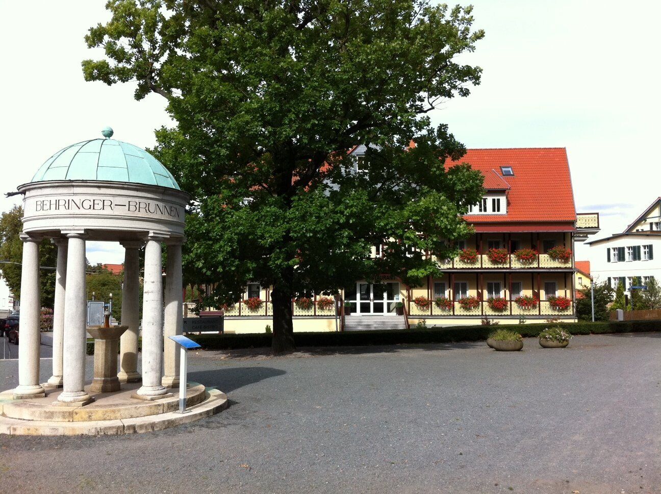 A gazebo with columns and a dome, surrounded by a paved area with a large tree and a building with balconies in the background.