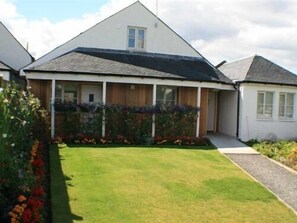A two-story house with a covered porch, a well-maintained lawn, and a garden with various plants and flowers. - Hillhead Farm Lets (Stirling)