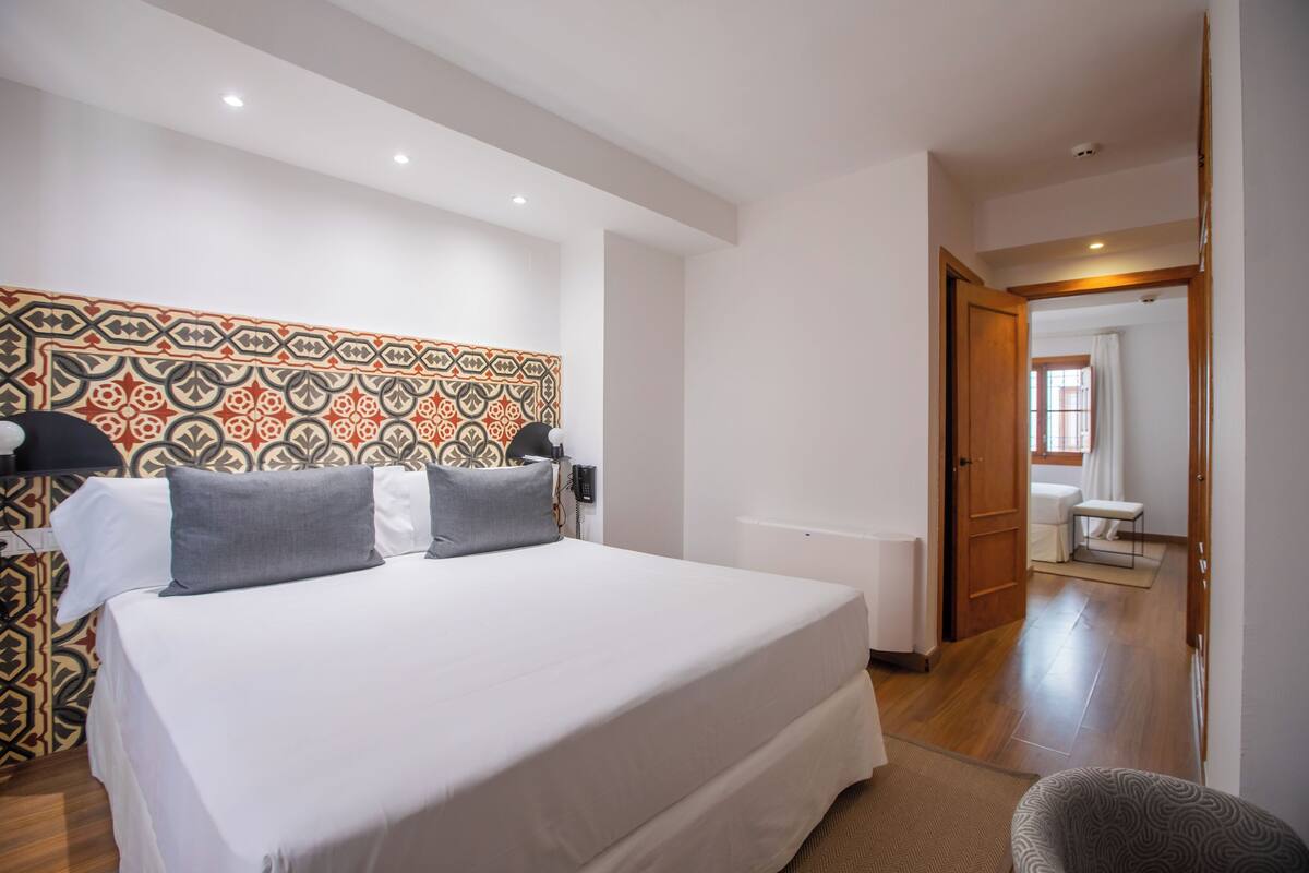 A hotel room with a large bed, a patterned headboard, a bedside table with a lamp, and a view into the bathroom.