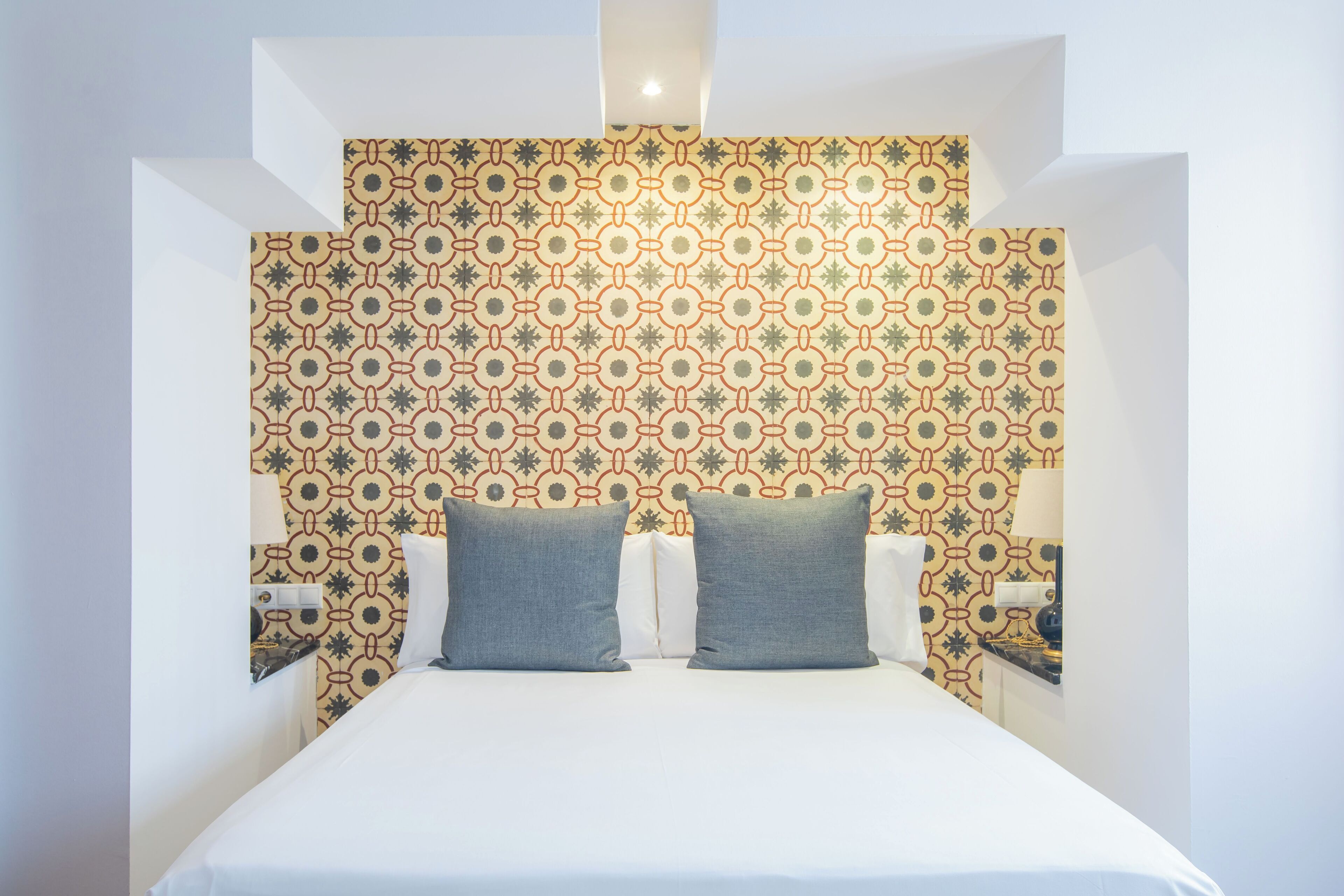 A neatly made bed with two pillows, a patterned wallpaper, and a bedside table with a lamp.