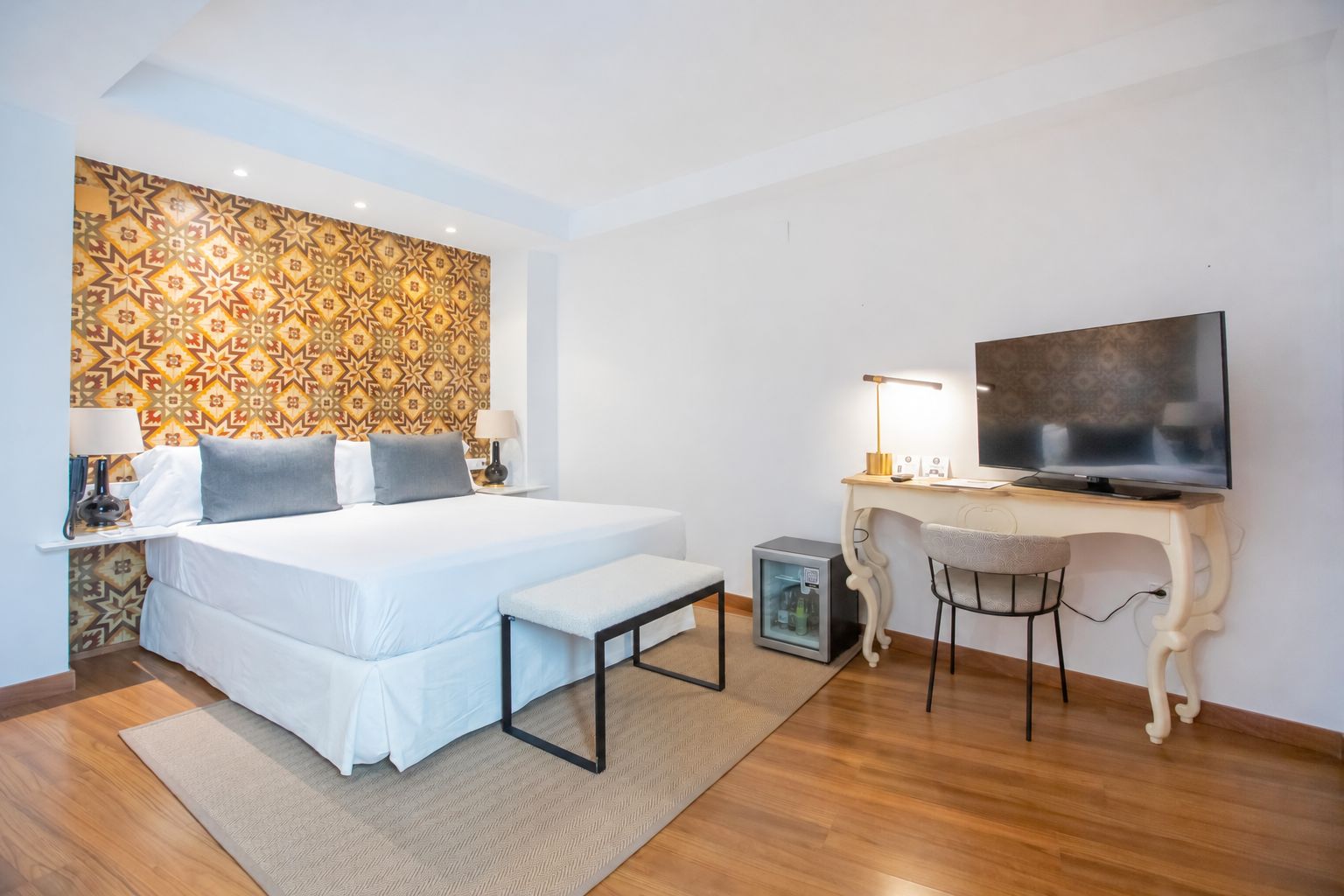 A hotel room with a bed, bedside table, lamp, desk, chair, television, and a small refrigerator.