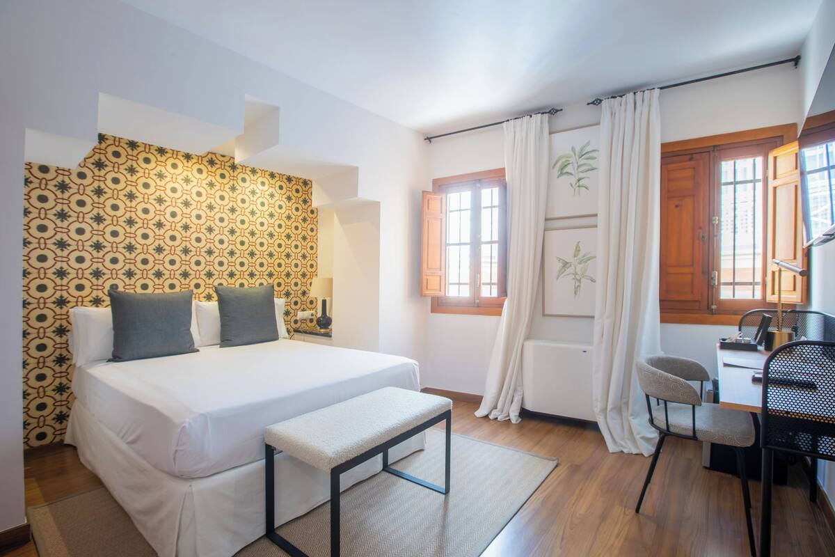 A bedroom with a bed, bench, desk, chair, and patterned wallpaper.