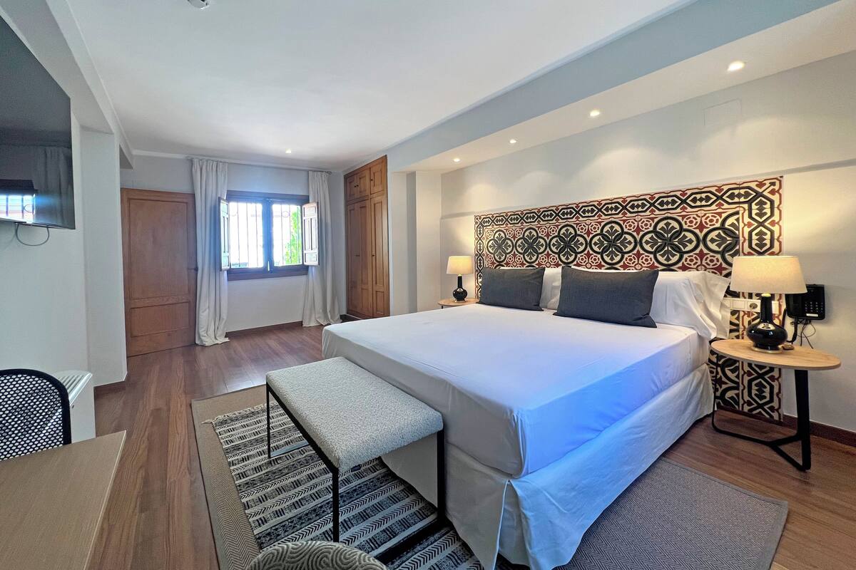 A modern hotel room with a large bed, two bedside tables with lamps, a bench, a TV, and a patterned headboard.