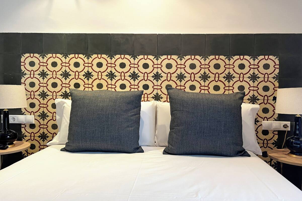 A neatly made bed with two pillows, a patterned wall behind, and bedside tables with lamps.