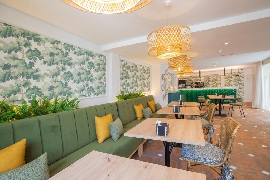 A modern restaurant with a green sofa, wooden tables, wicker chairs, and a patterned wall.