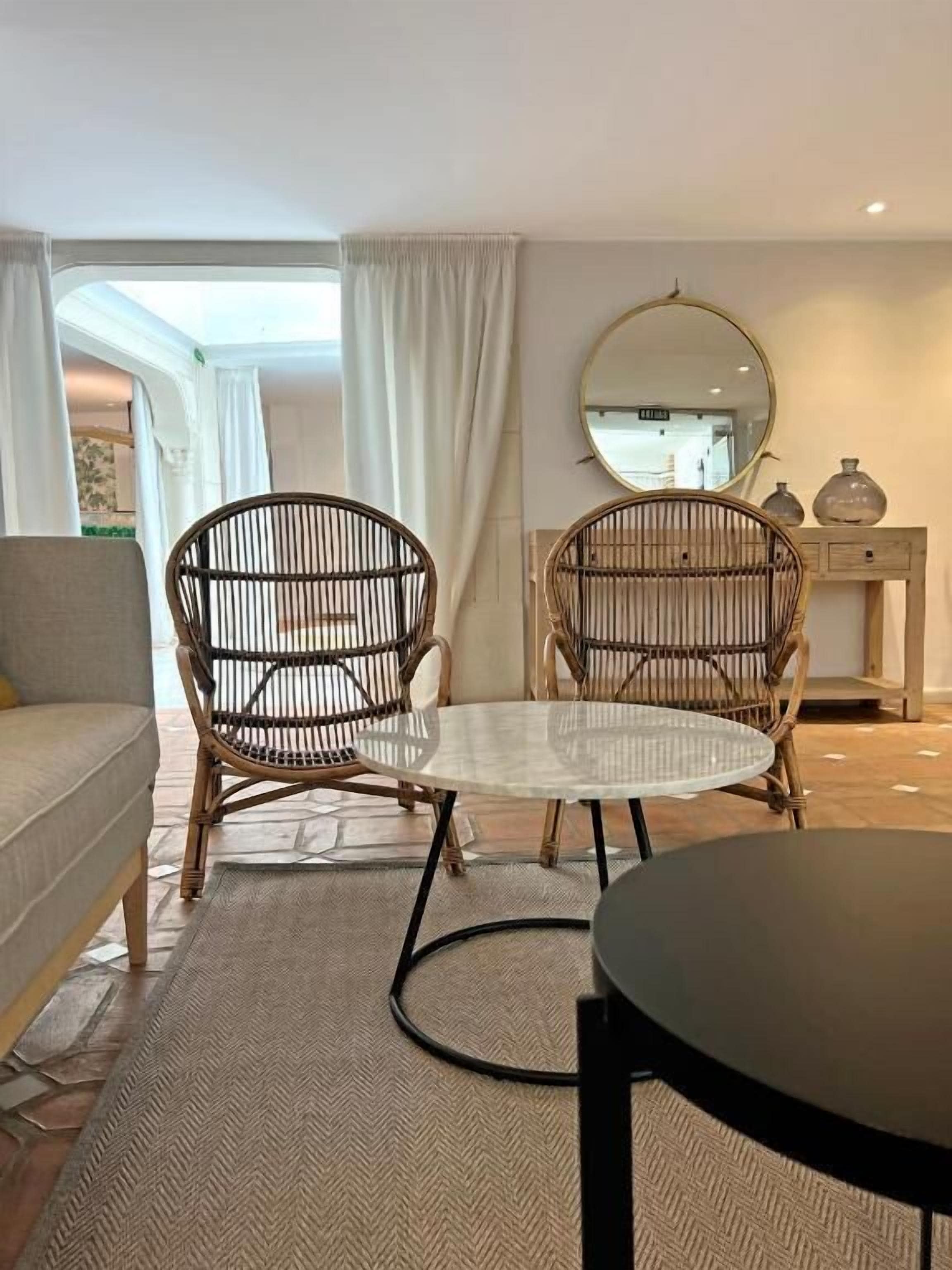 Een kamer met een bank, twee rieten stoelen, een ronde tafel, een spiegel en een console met decoratieve objecten.