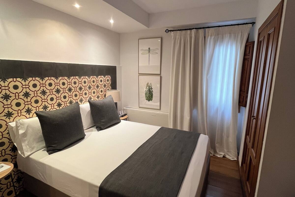 A hotel room with a large bed, a headboard with a geometric pattern, a bedside table with a lamp, and a window with white curtains.