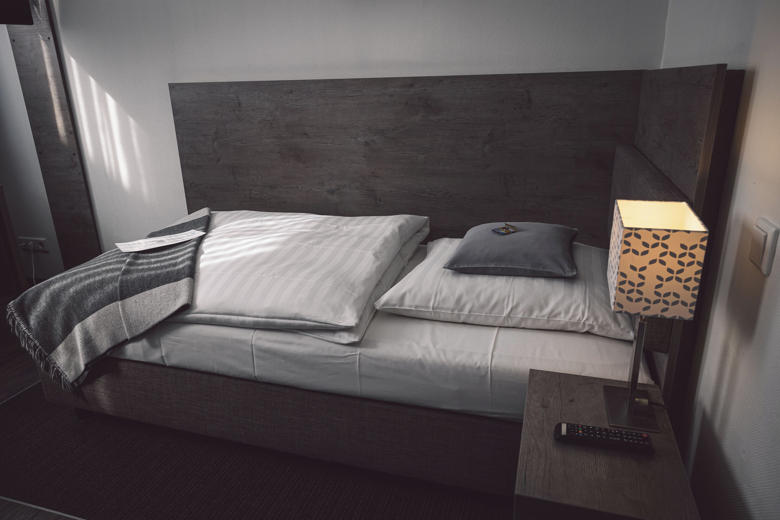 Uma cama com cabeceira de madeira, um controle remoto sobre uma mesa de cabeceira e um abajur estampado.