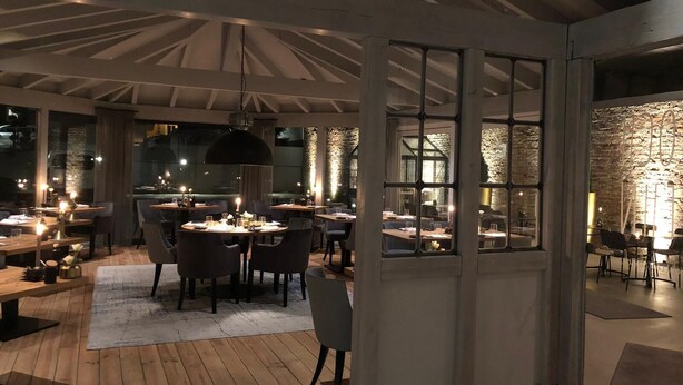 A modern dining area with wooden flooring, round tables, and chairs. There are large windows with a stone wall visible through them. - Hotel Alte Redaktion Superior (Gevelsberg)