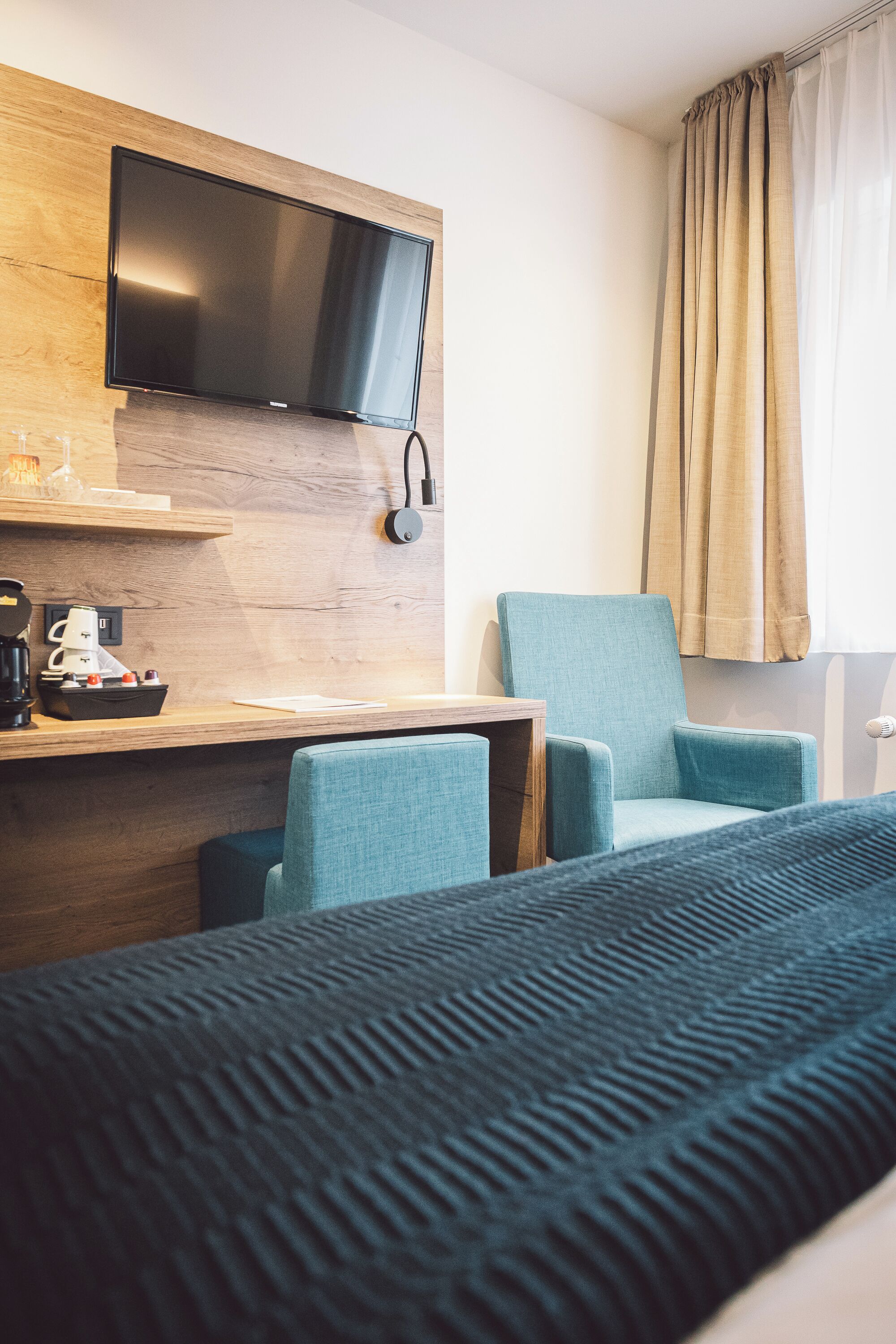 A hotel room with a bed, a desk with a coffee maker, a television mounted on a wooden wall, and a chair.