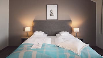 Una cama doble con una colcha blanca, dos mesitas de noche con lámparas, un cuadro enmarcado en la pared y un muro con textura.