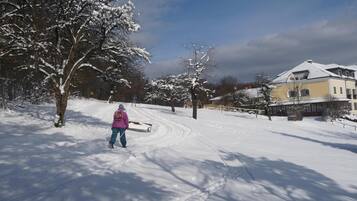 Schneebedeckte Landschaft mit einer Person, die auf einem Weg unterwegs ist, Bäume und einem Gebäude im Hintergrund.