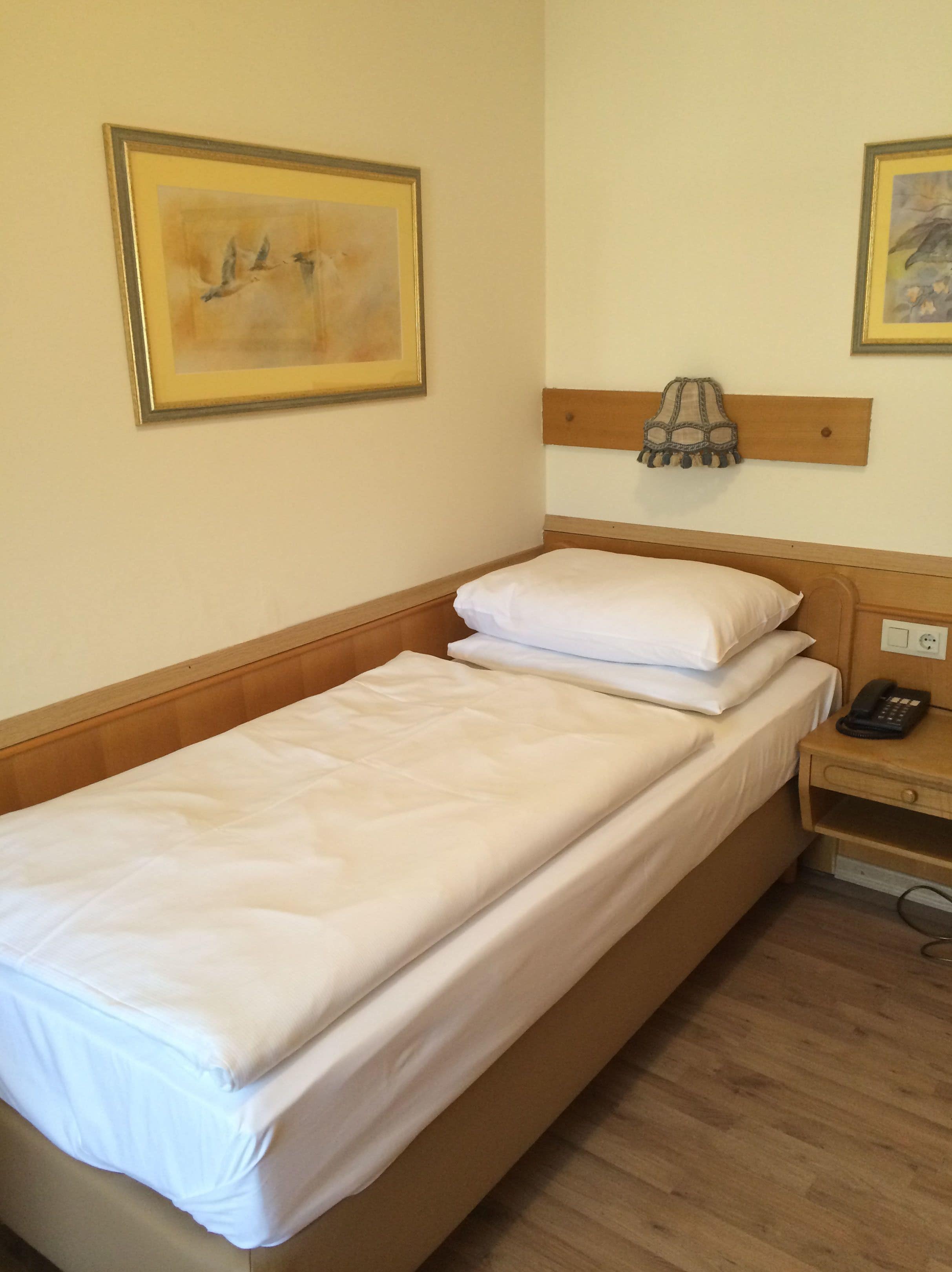 Una cama individual con sábanas blancas, cabecera de madera, mesita de noche con lámpara y teléfono.
