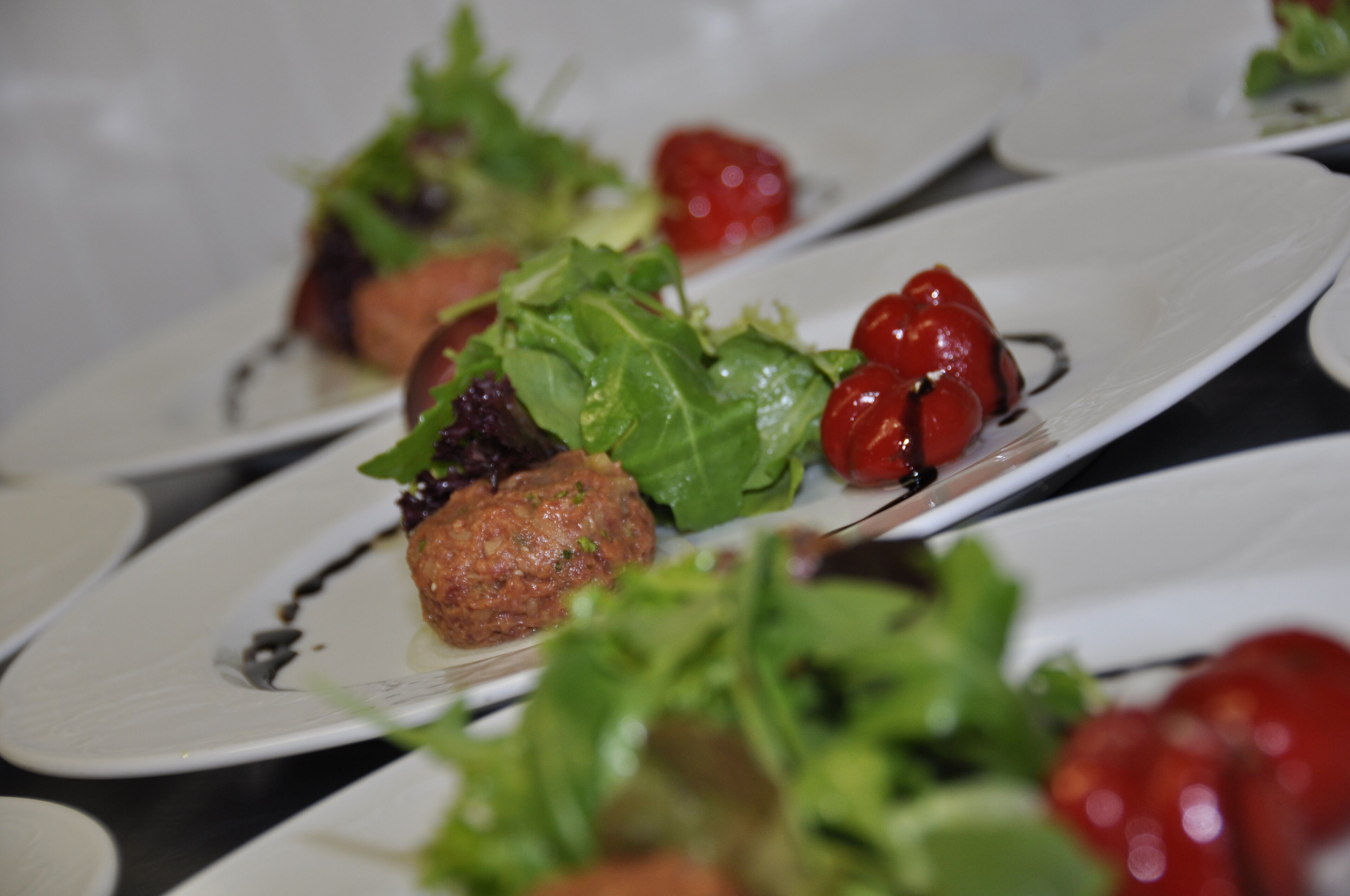 A series of white plates with food items, including a meat patty, greens, and cherry tomatoes.