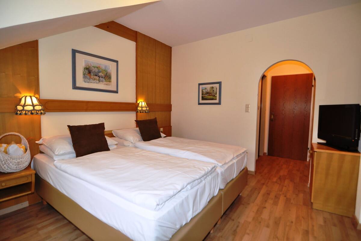 A hotel room with two beds, wooden headboards, wall-mounted lamps, framed pictures, a television on a wooden stand, and a basket on the nightstand.