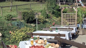 Een buiten eetgedeelte met houten banken en tafels, omgeven door groen en een houten pergola.