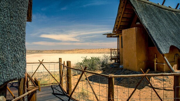 A desert landscape with a wooden structure featuring a thatched roof and a wooden deck leading to a fenced area. - Xaus Lodge (Mier)