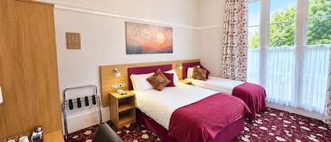 A hotel room with two beds, a desk with a kettle, a chair, a painting on the wall, and a large window with curtains.