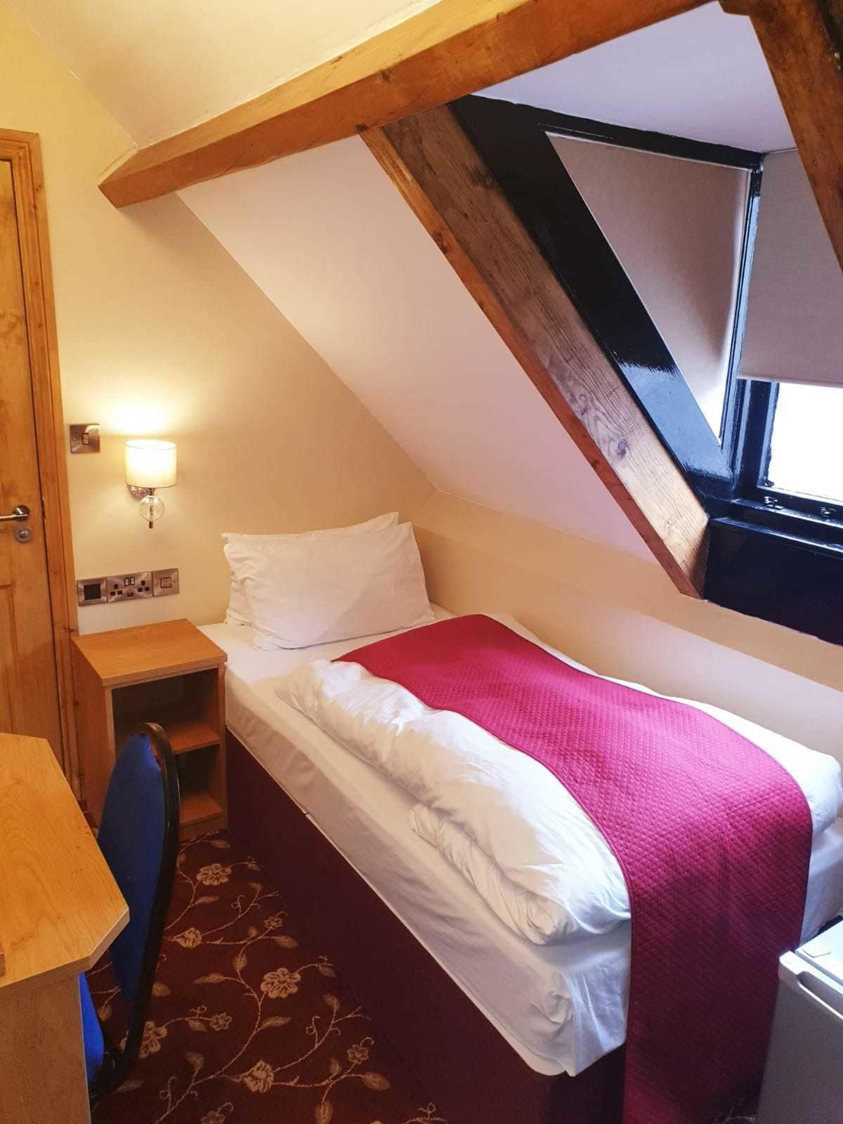 Une petite pièce accueillante avec un plafond en pente, un lit simple avec une literie blanche et une couverture rouge, un bureau avec une chaise, une lampe fixée au mur et une fenêtre.