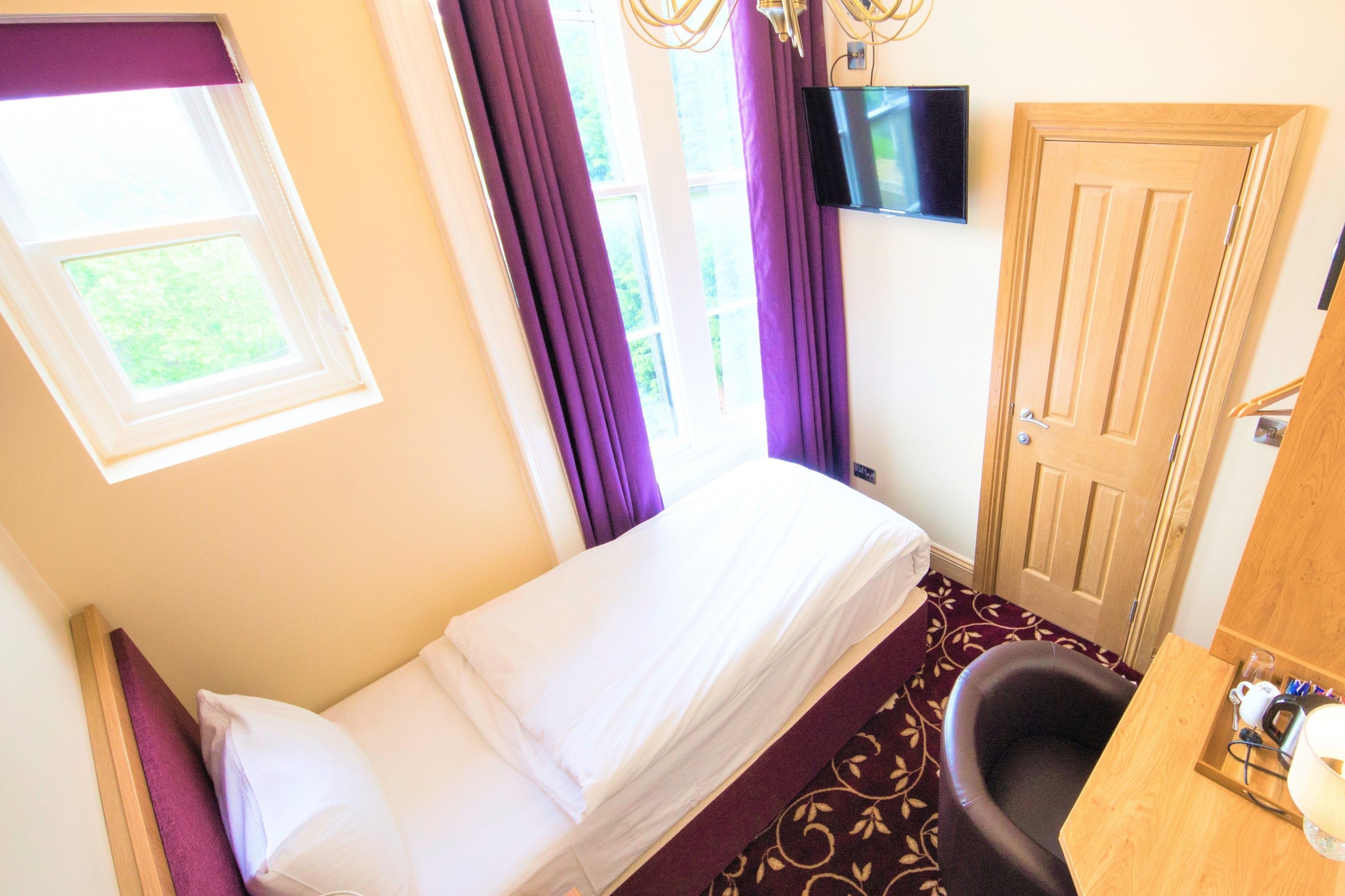 A single bed with white linens, a window with purple curtains, a television mounted on the wall, and a wooden door.