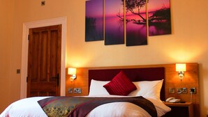 A hotel room with a large bed, a wooden door, and three wall-mounted paintings depicting a sunset over water. - Edgerton Hotel (Huddersfield)