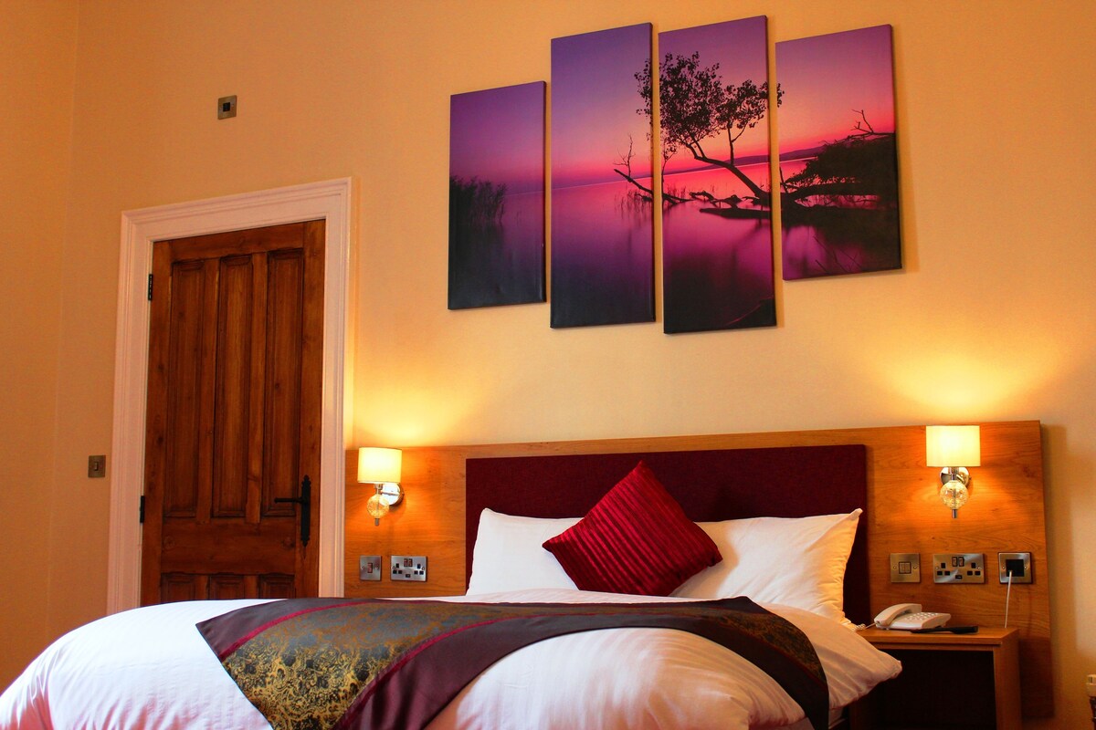 A hotel room with a large bed, a wooden door, and three wall-mounted paintings depicting a sunset over water.
