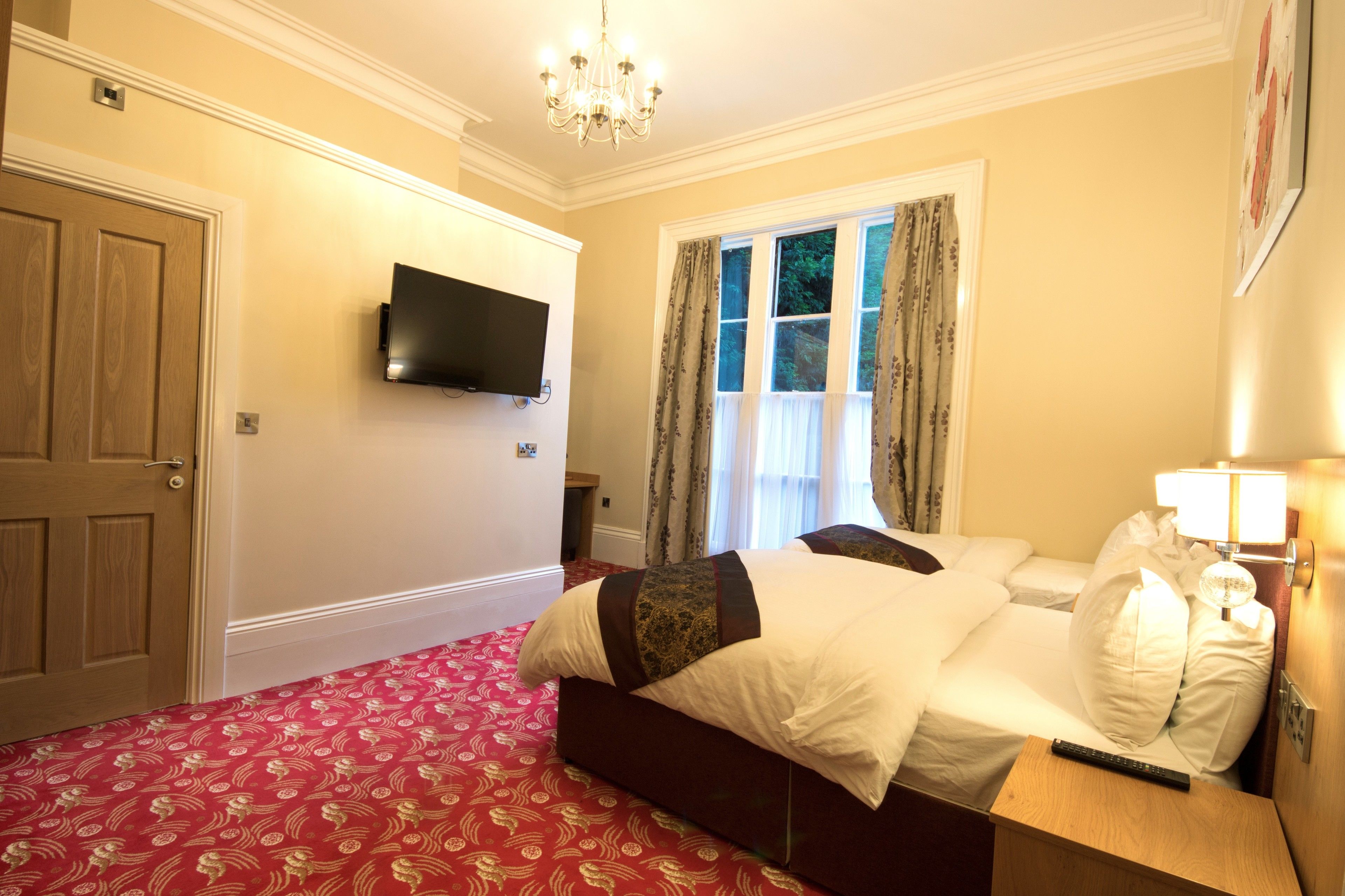 A hotel room with a large bed, a television mounted on the wall, a window with curtains, and a chandelier.