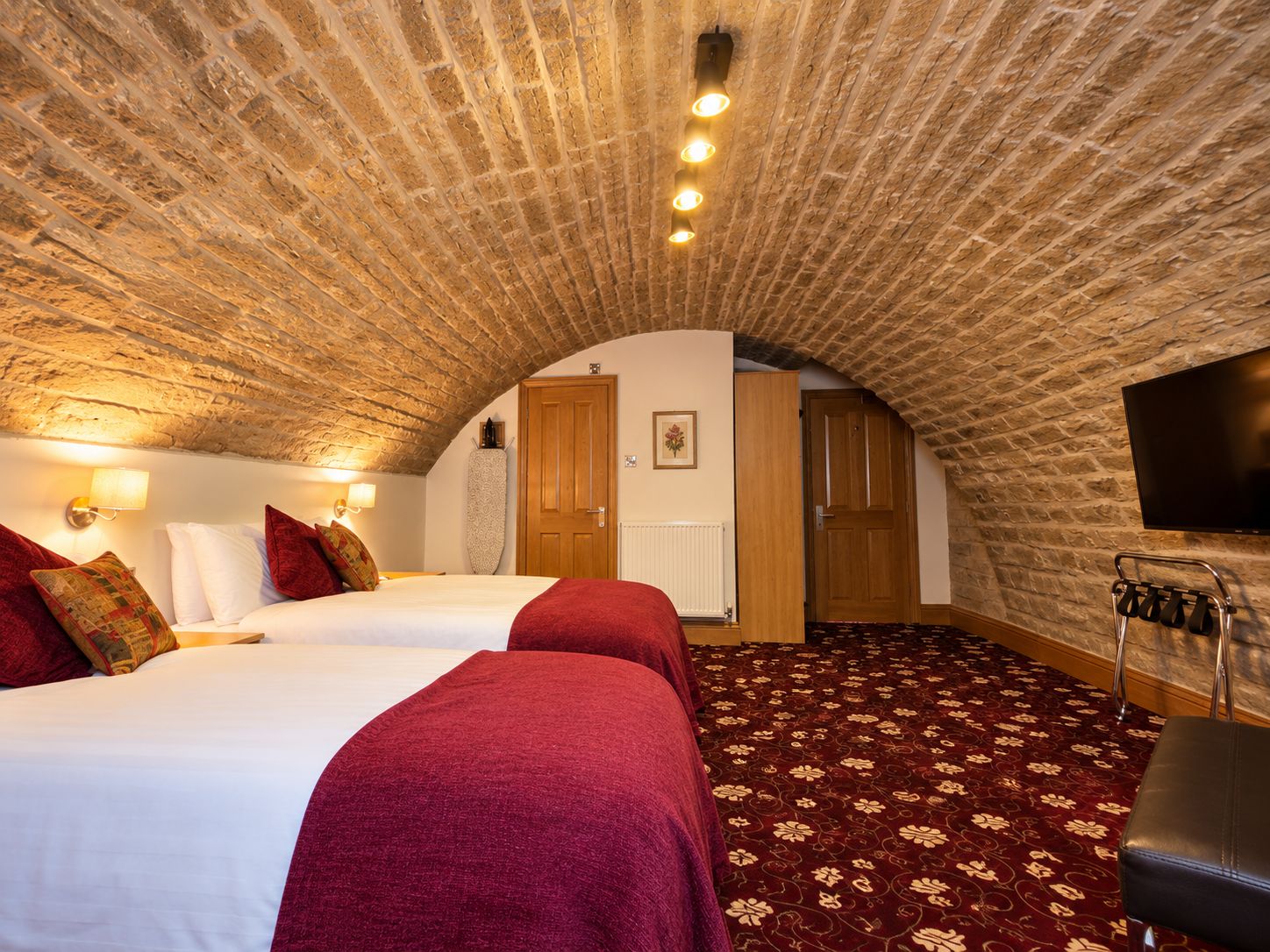 A room with a vaulted ceiling, two beds with red bedding, a patterned carpet, a television mounted on the wall, and a wooden door.