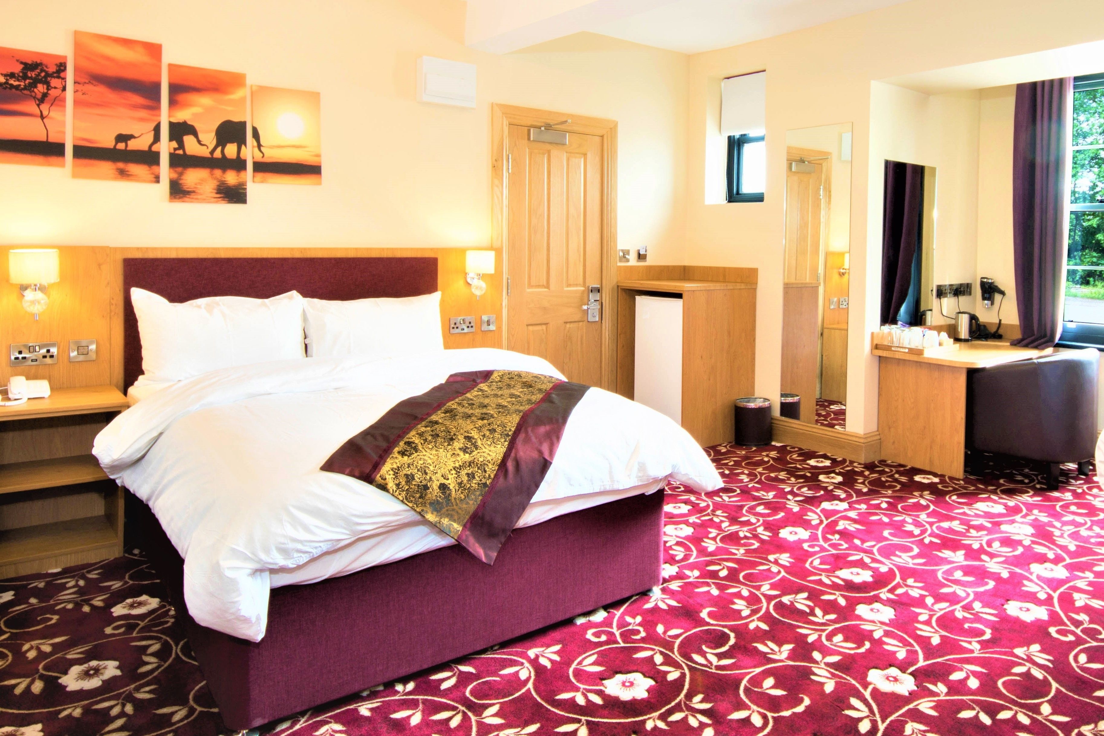 A hotel room with a large bed, a wooden headboard, a mirror, a vanity with a sink, and a window with curtains.