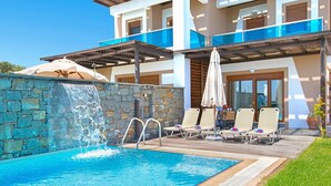 Una piscina con cascada, rodeada de sillones y sombrillas, con un muro de piedra y un edificio de varios pisos al fondo.