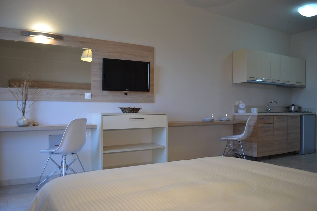 Moderni hotellihuone, jossa on sänky, työpöytä tuolilla, televisio ja pieni keittiönurkkaus.
