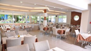 A spacious restaurant with multiple tables and chairs, large windows offering a view of the outdoors, and a modern interior design. - Argiro Village (Rhodes)