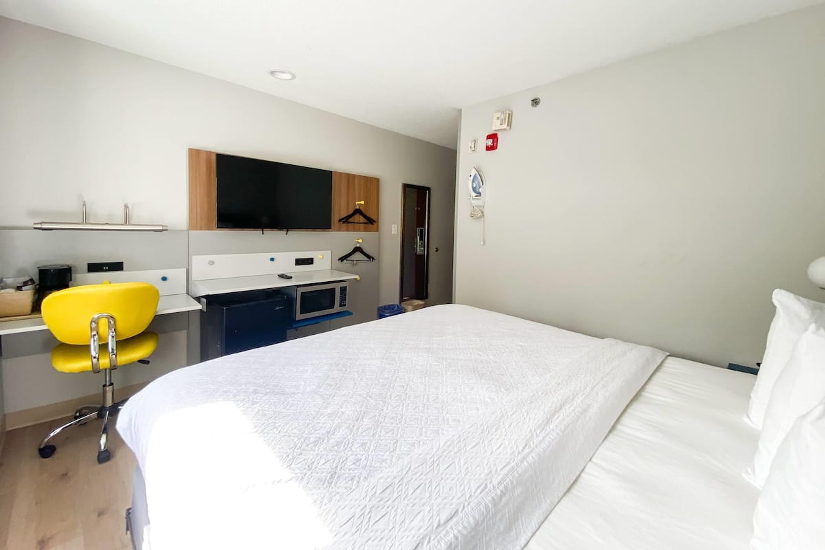 A hotel room with a bed, a desk with a chair, a microwave, and a TV mounted on the wall.