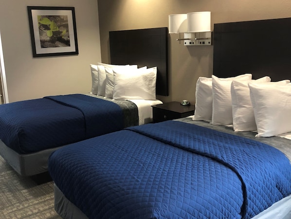 A hotel room with two beds, each with blue bedding, a nightstand with a lamp, and a framed picture on the wall.