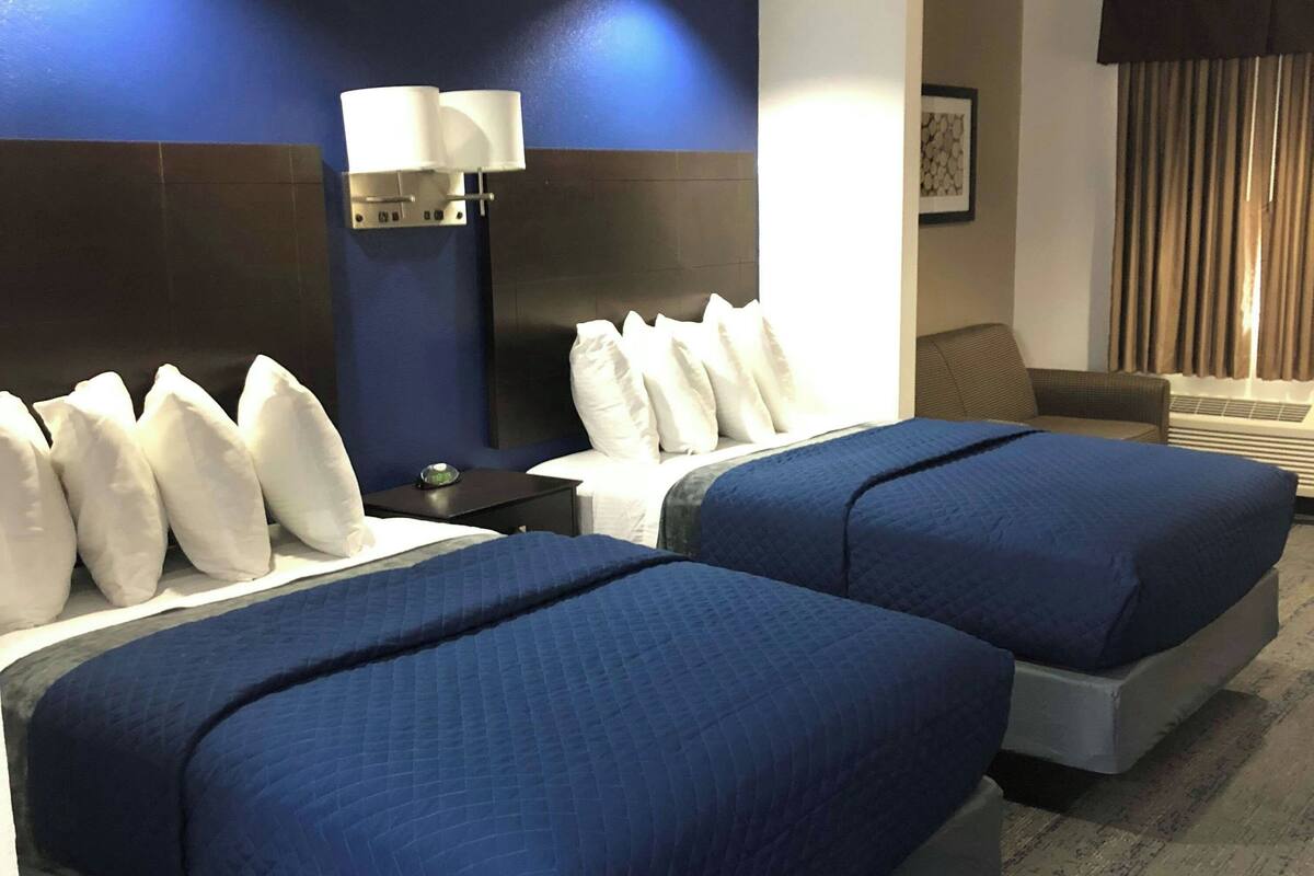 Ein Hotelzimmer mit zwei Betten, einem Nachttisch mit Lampe, einem Sofa und einem an der Wand hängenden Kunstwerk.
