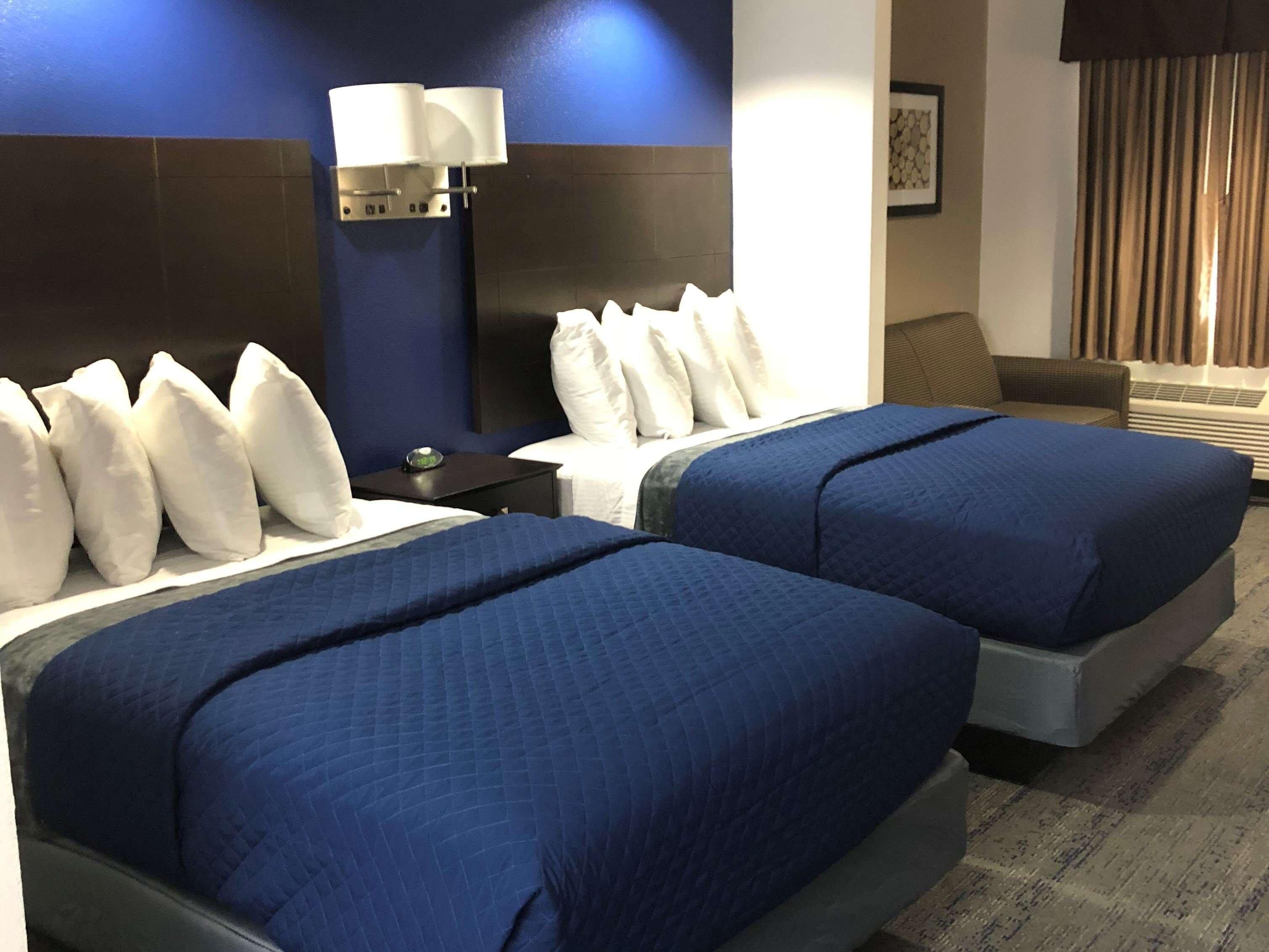 A hotel room with two beds, a nightstand with a lamp, a sofa, and a window with curtains.