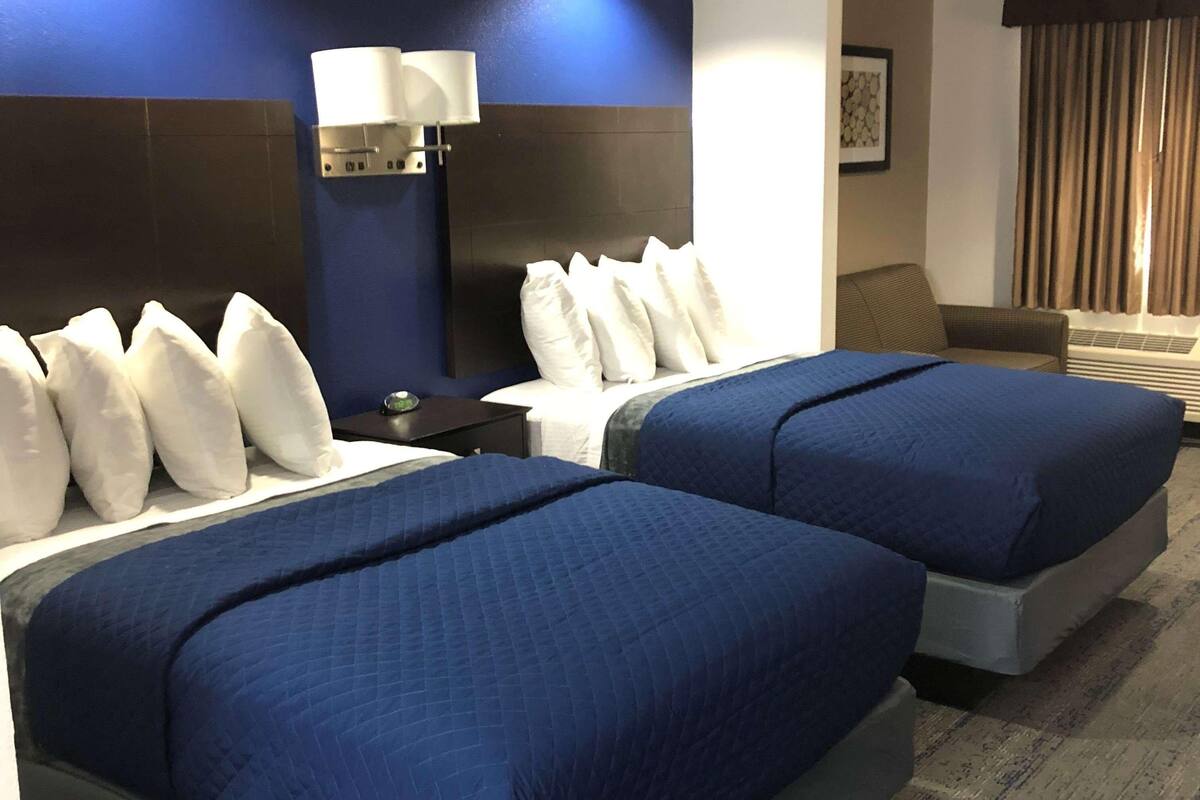 Ein Hotelzimmer mit zwei Betten, einem Nachttisch mit Lampe, einem Sofa und einem Fenster mit Vorhängen.