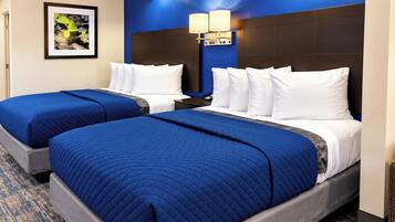A hotel room with two beds, each with a blue quilted bedspread and white pillows. There is a framed picture on the wall and a nightstand with a lamp between the beds.