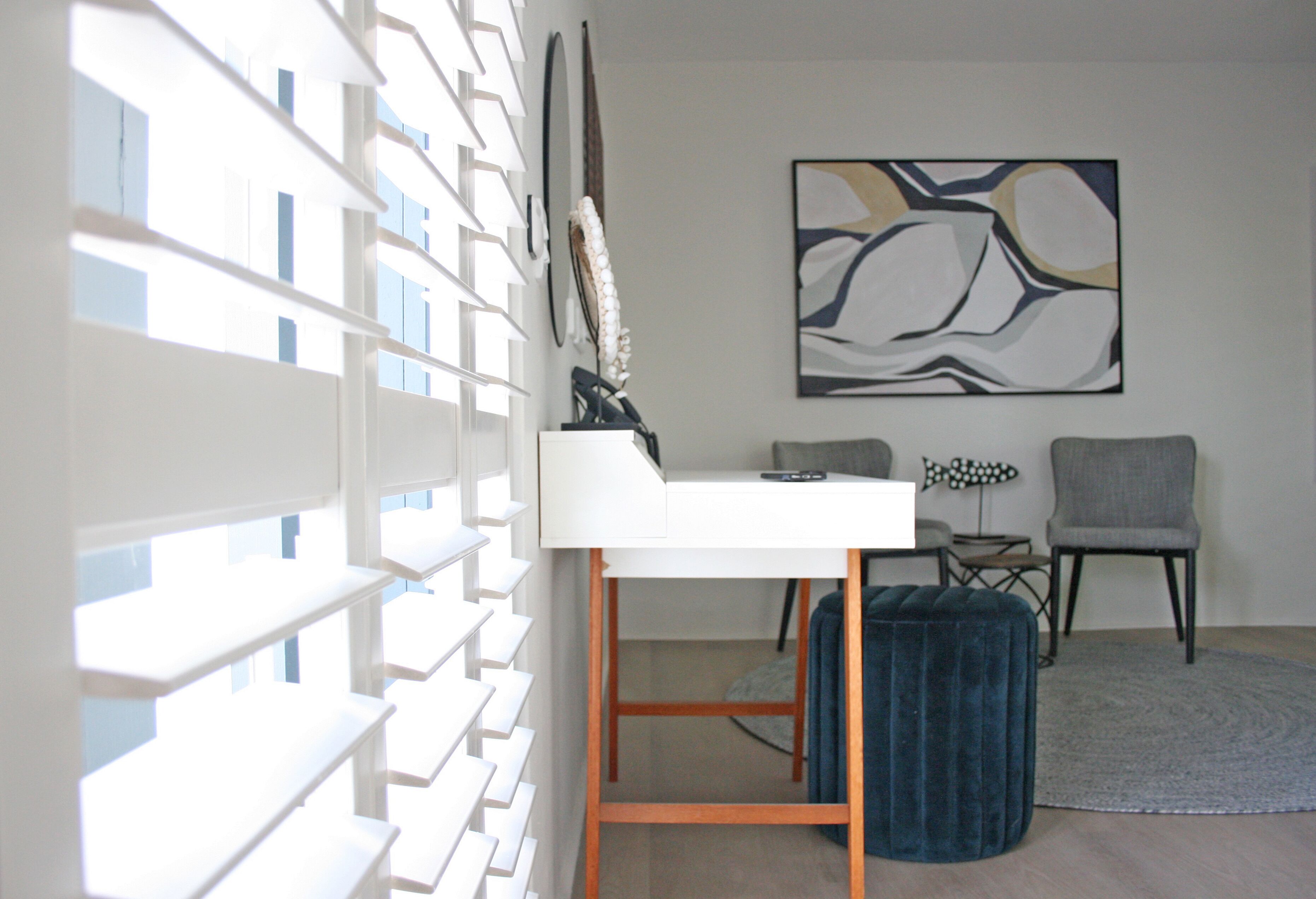 A modern room with a white desk, a grey chair, a blue ottoman, and a large abstract painting on the wall.