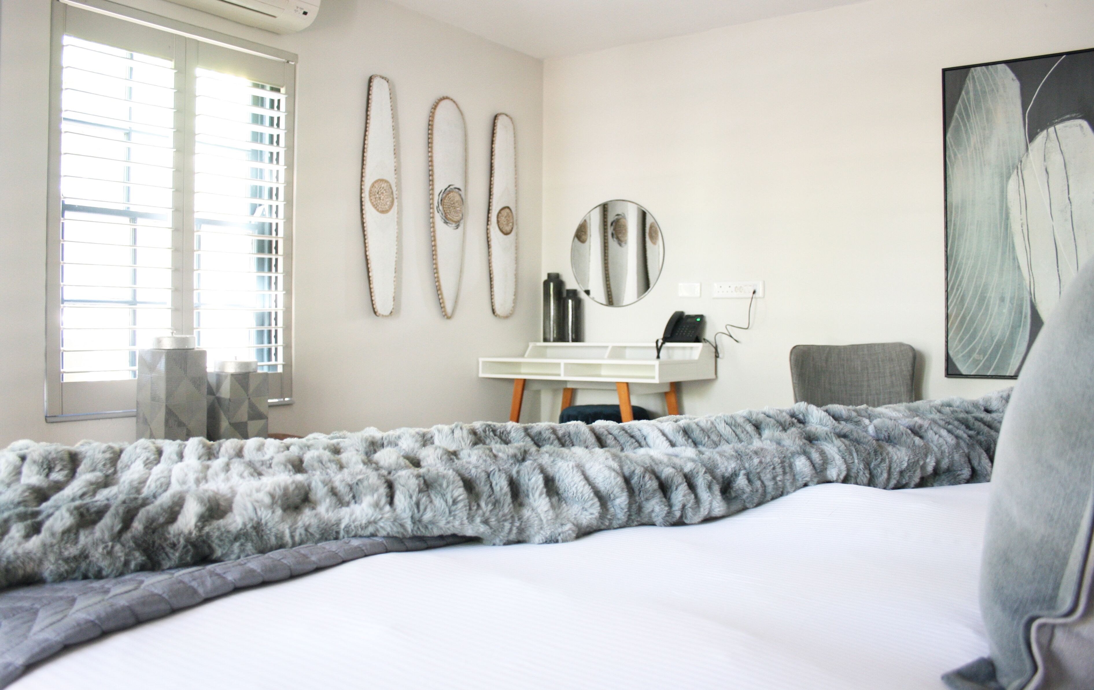 Ein modernes Hotelzimmer mit einem großen Bett, einem Schreibtisch und einem Sessel. An der Wand befinden sich dekorative Elemente und ein Fenster mit Jalousien.