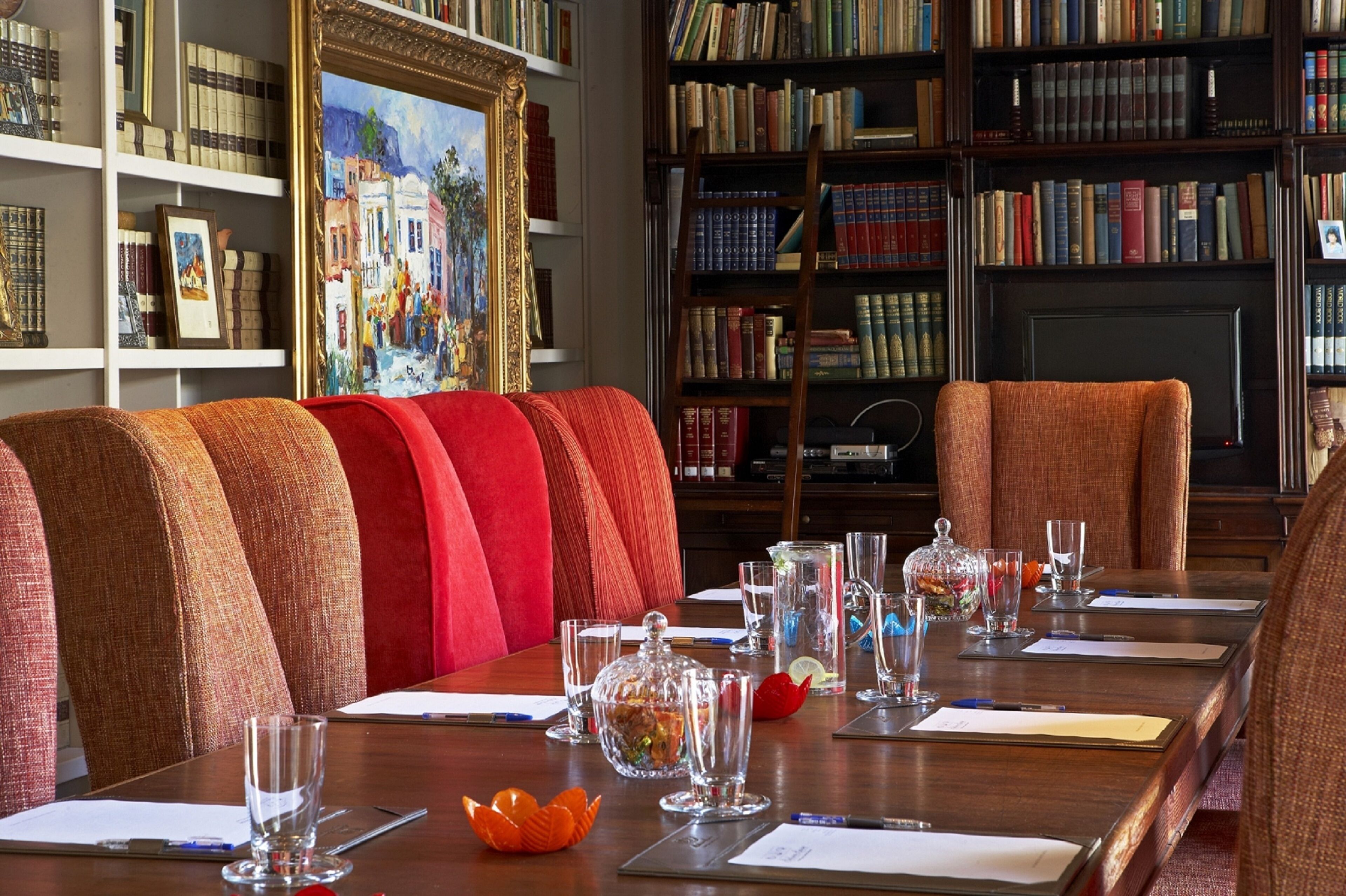 A conference room with a long table, red upholstered chairs, and a bookshelf filled with books.