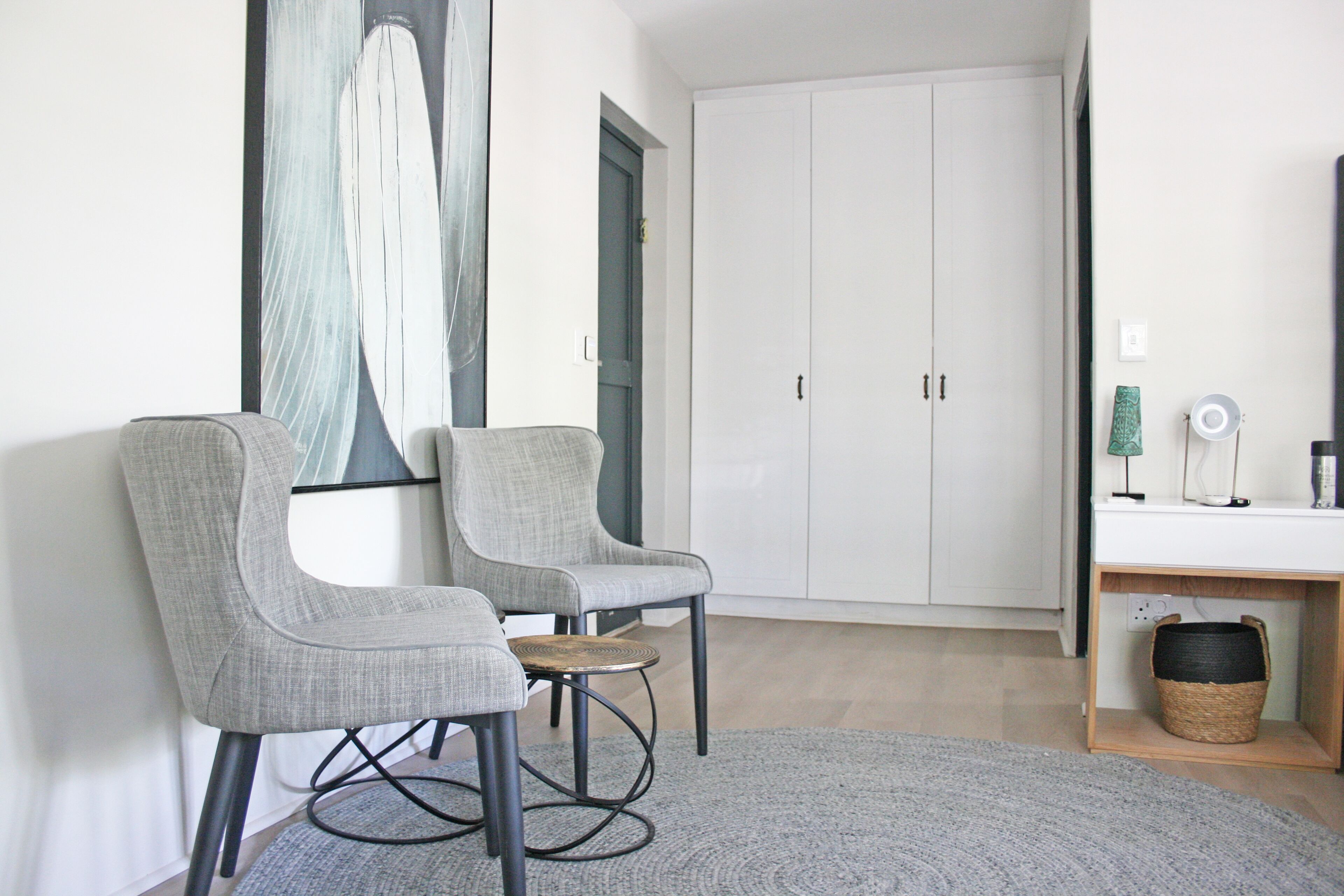 Quarto moderno e bem iluminado com duas poltronas cinzas, uma mesa redonda pequena, um tapete cinza, um guarda-roupa branco e uma luminária fixada na parede.