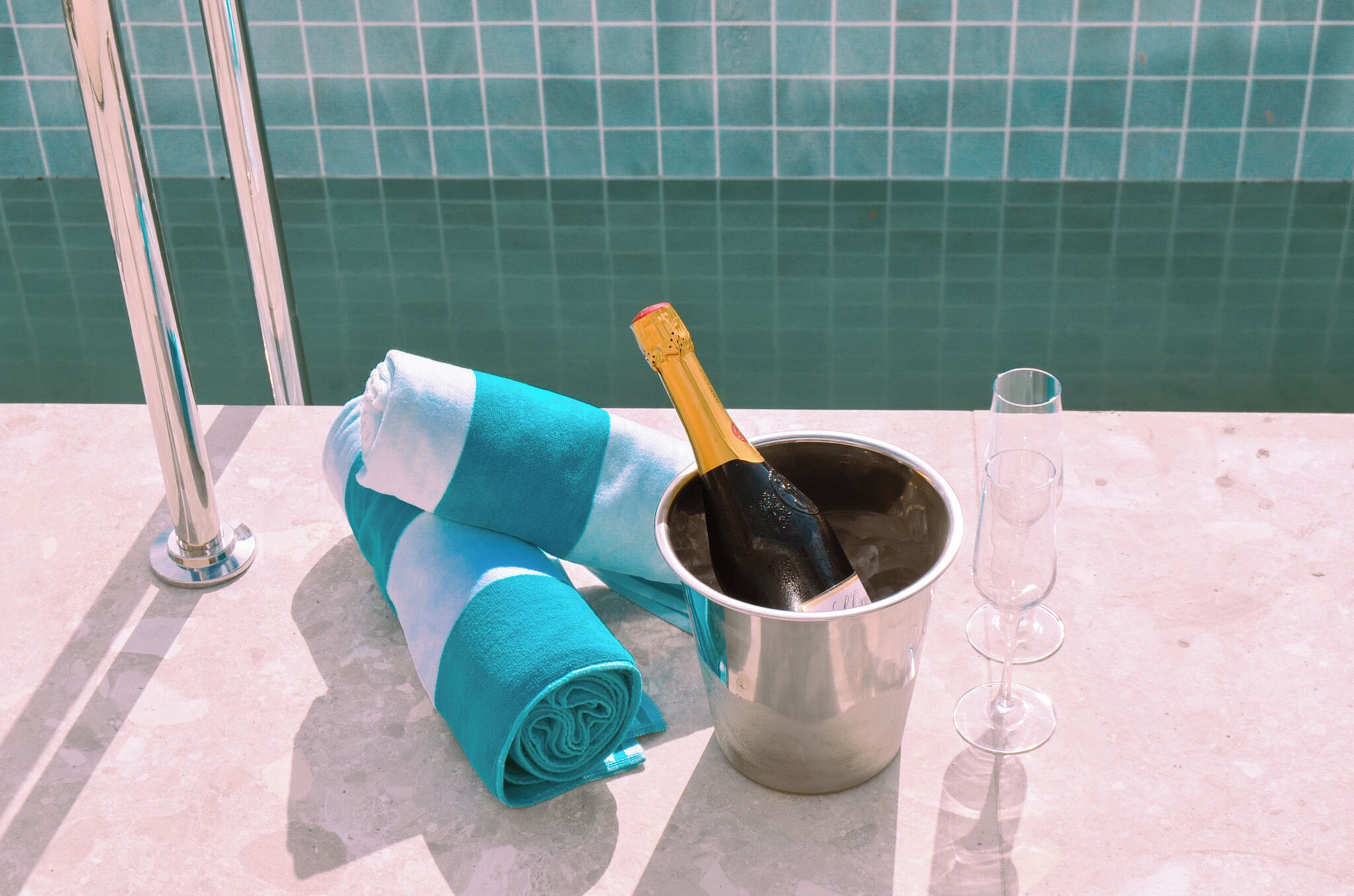 A metal bucket with a bottle of champagne and a glass on a poolside surface.