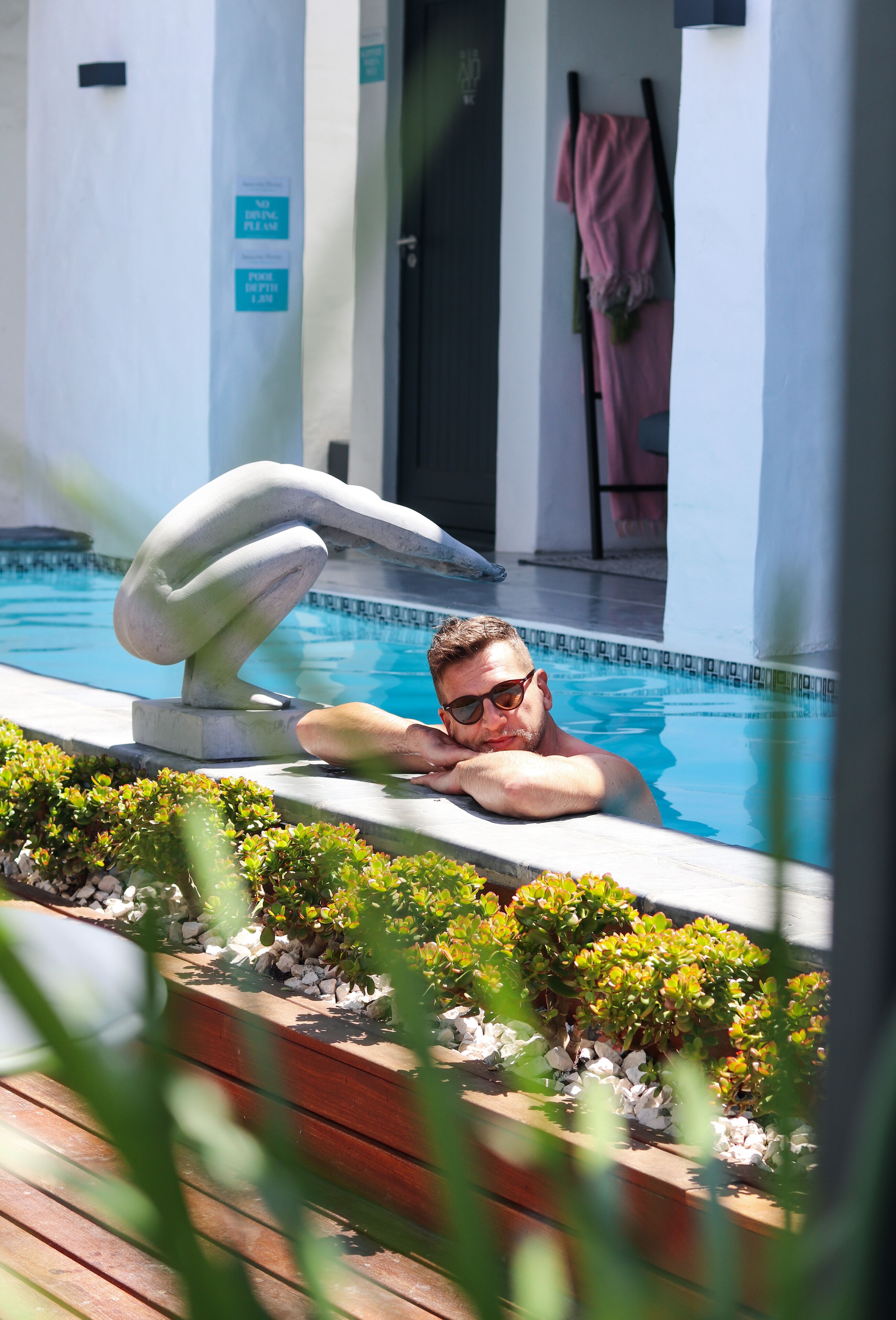 A man relaxing in a swimming pool with a stone sculpture and a towel rack in the background.